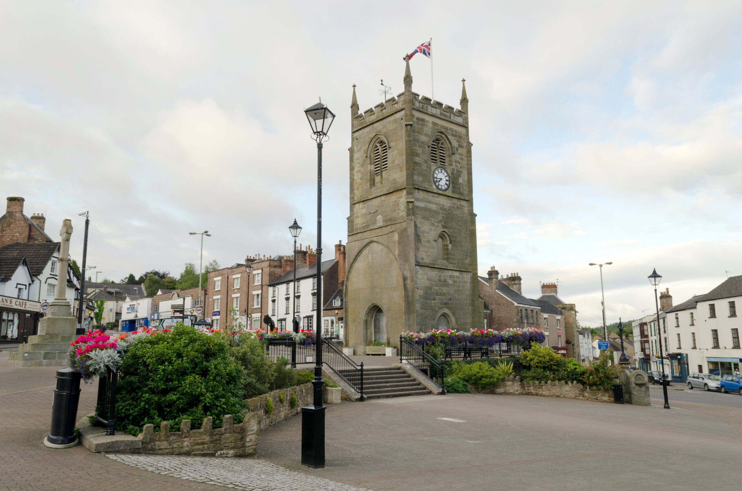 A stone clock tower with a Union Jack flag sits in a town square, surrounded by flower beds and lampposts. Historic buildings, partly hidden by commercial scaffolding, are visible in the background under a cloudy sky.