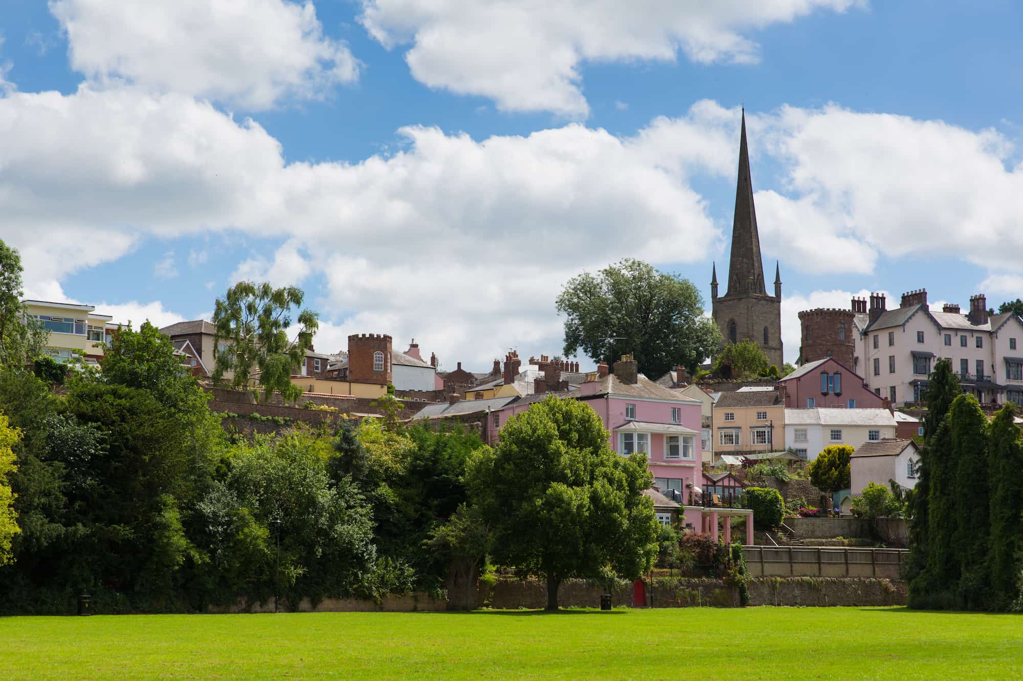 A scenic view of a town with colorful houses, lush green trees, and a tall church spire rising above the rooftops under a partly cloudy blue sky.