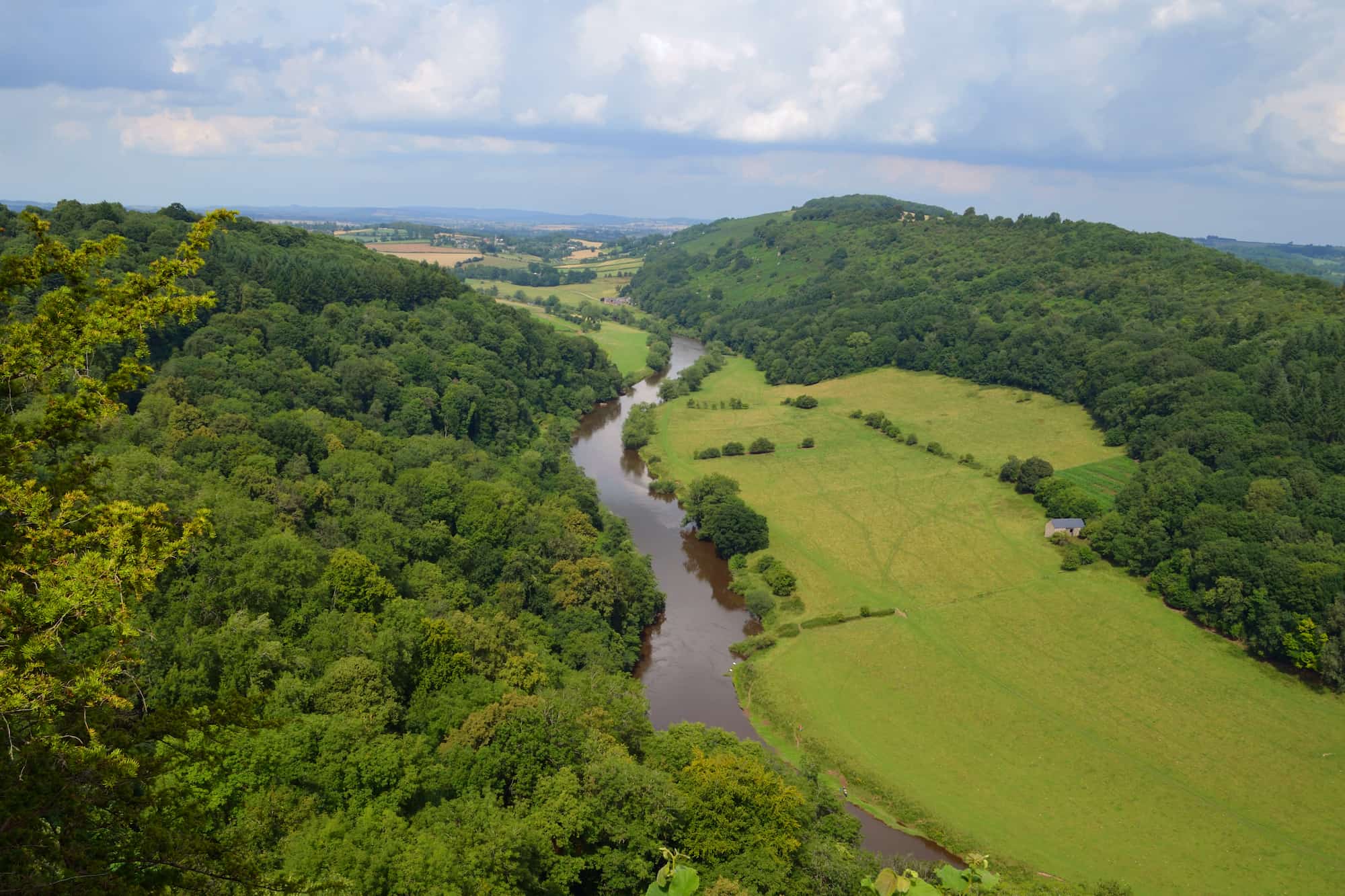 A river winds through a lush green valley, bordered by dense trees on one side and open grassy fields on the other, with rolling hills and a cloudy sky in the background.