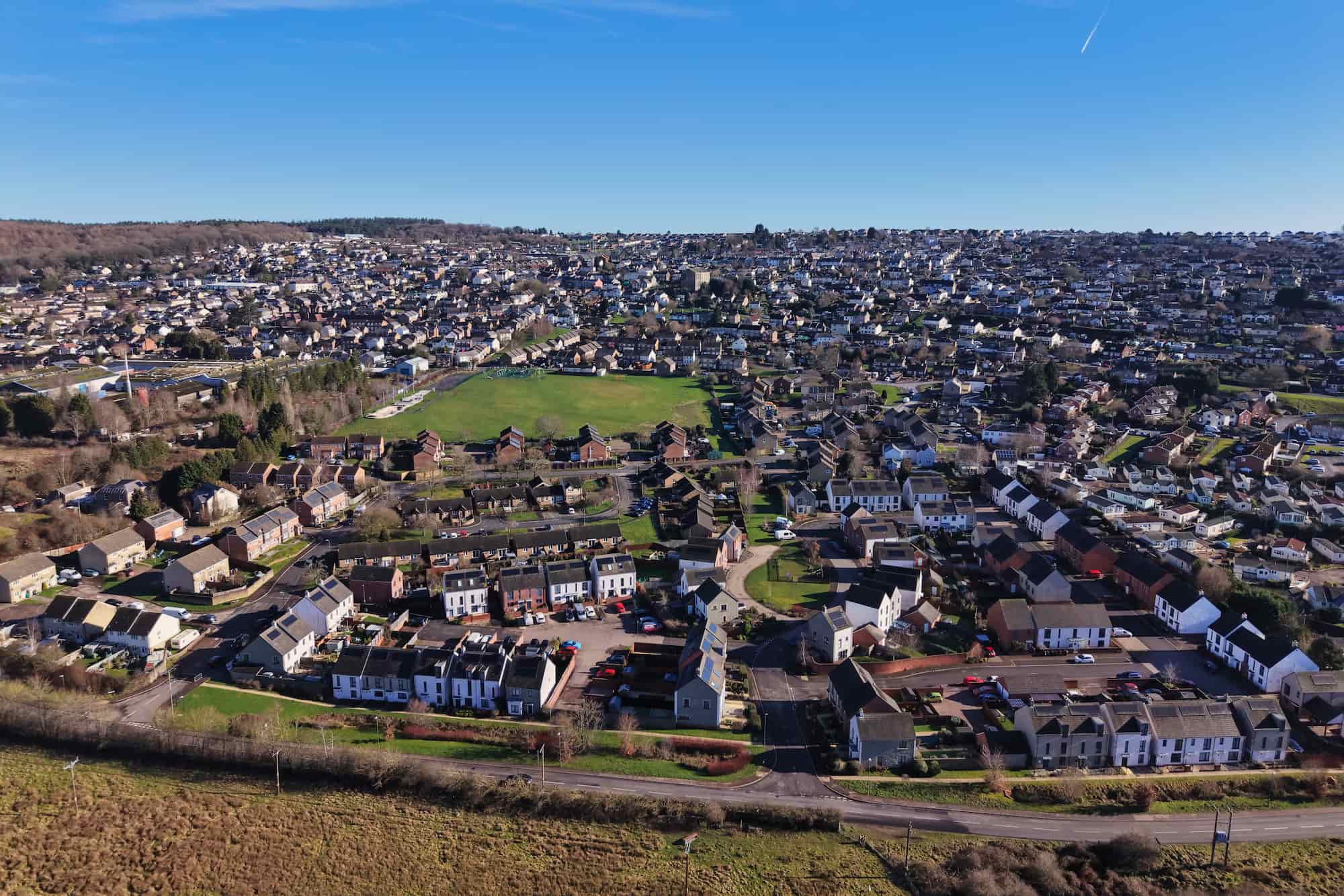 Aerial view of a suburban town with rows of houses, green spaces, and roads under a clear blue sky on a sunny day.