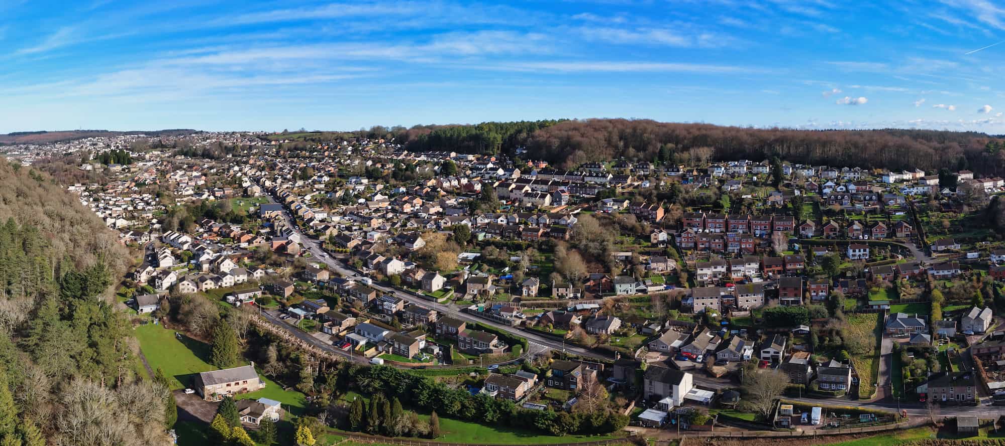 Aerial view of a small town with houses, roads, and buildings surrounded by trees, hills, and greenery under a bright blue sky with light clouds.