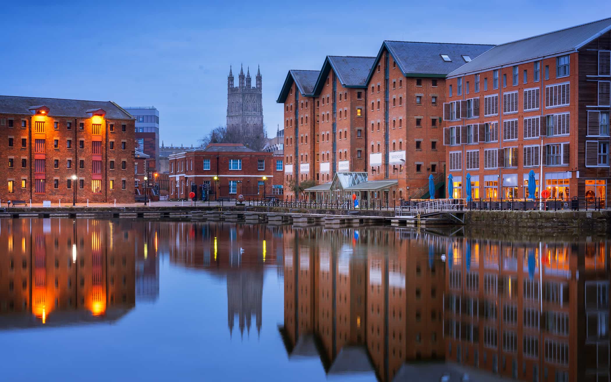 Historic brick warehouses and modern buildings line a calm waterfront at dusk, with warm lights reflected in the water. A tall cathedral tower rises in the background under a blue sky.