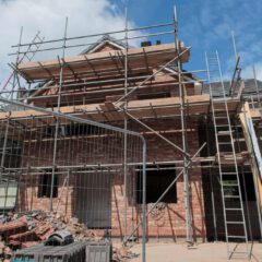 A brick house under construction with metal scaffolding on all sides, ladders leaning against the structure, and piles of building materials and debris in the foreground under a partly cloudy sky.