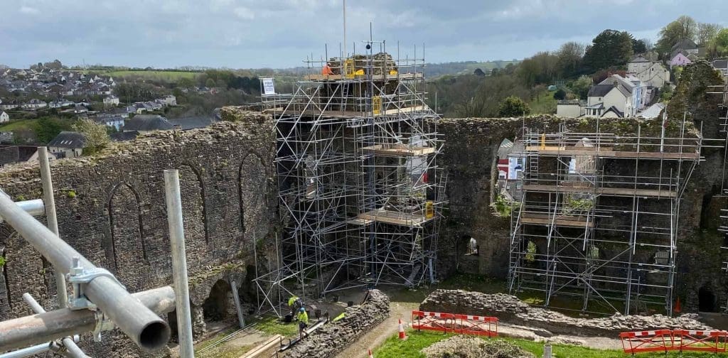 Scaffolding covers parts of an old stone castle ruin under restoration, with workers on site. Surrounding villages and green hills are visible in the background under a cloudy sky.
