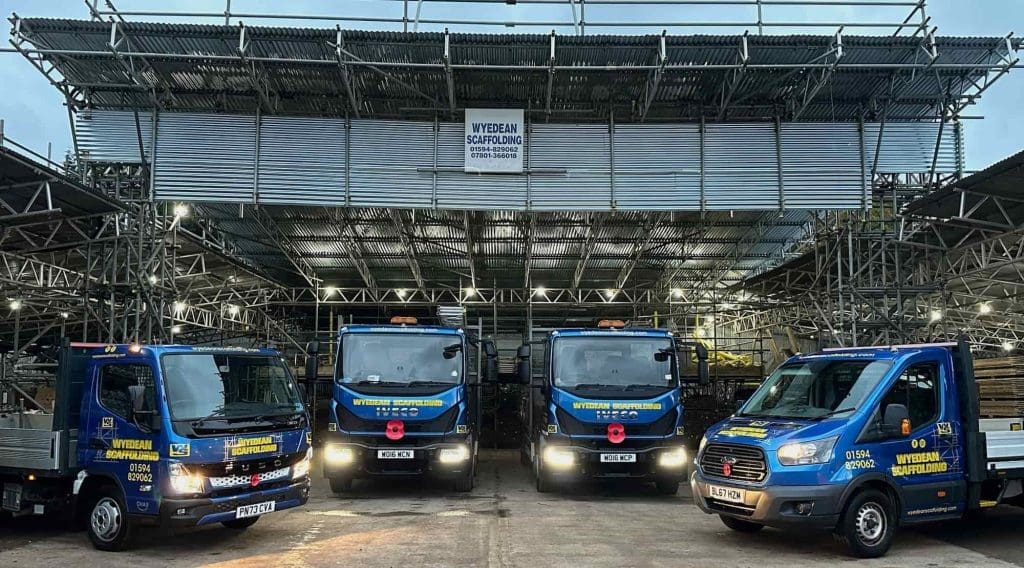 Four blue company trucks with yellow lettering are parked in front of a large scaffolding structure at dusk. The trucks display "Wyreban Scaffolding" and contact details on the doors.