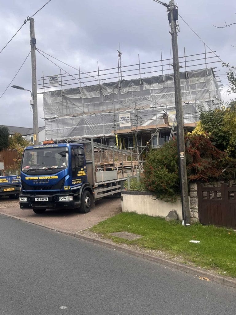 A house under construction is covered in scaffolding and white sheeting. A blue flatbed truck with “Skyline Solutions” branding is parked in front. The sky is cloudy and there are trees and a garden nearby.