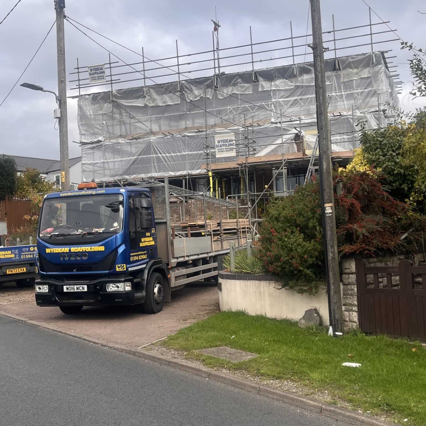 A house under construction is covered in scaffolding and white sheeting. A blue flatbed truck with “Skyline Solutions” branding is parked in front. The sky is cloudy and there are trees and a garden nearby.